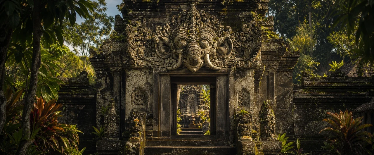 A weathered stone Karang Asti carving of a fierce elephant face crowns a Balinese temple doorway, moss-covered and framed by tropical foliage, with a second gateway visible through the passage beyond.