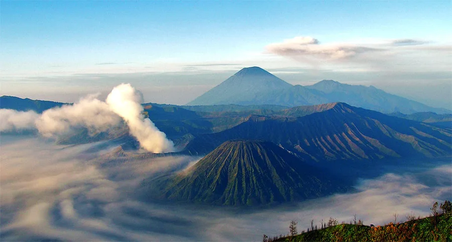 Mount Bromo's volcanic crater releasing steam, seen from above the clouds with Mount Semeru in the distance.