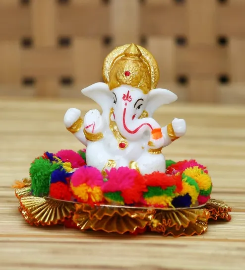 A golden Ganesh idol holding modaks, seated before a blurred backdrop of glowing string lights.