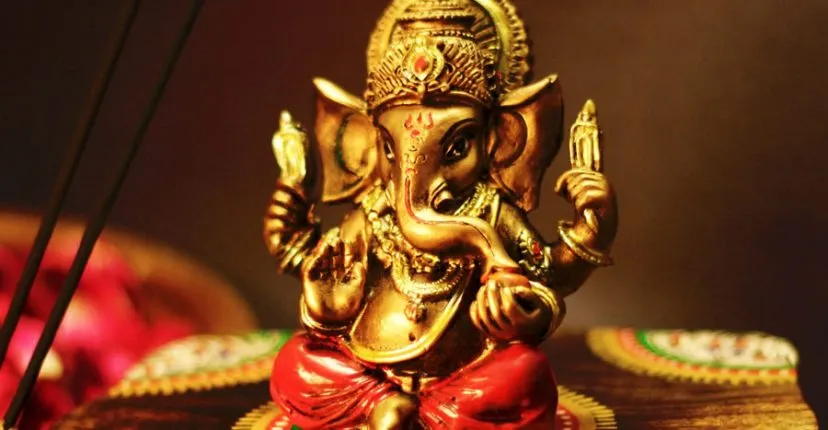 A close-up of a golden Ganesh idol lit by candlelight, with an incense stick burning beside it.