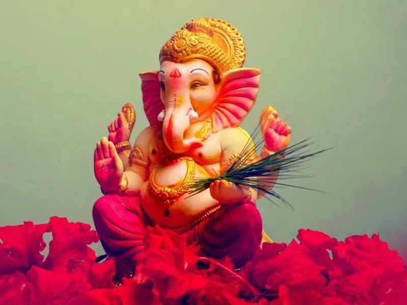 A golden Ganesh idol holding a sprig of durva grass, seated amid red flowers.