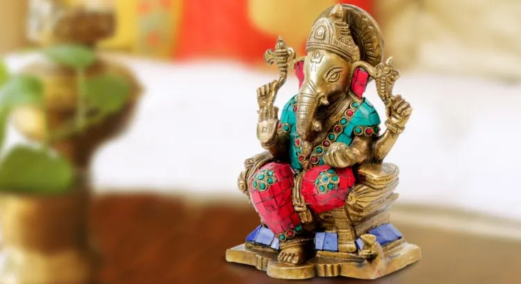 A small brass Ganesh idol inlaid with turquoise and red stones, seated on a red cushion beside a potted plant.